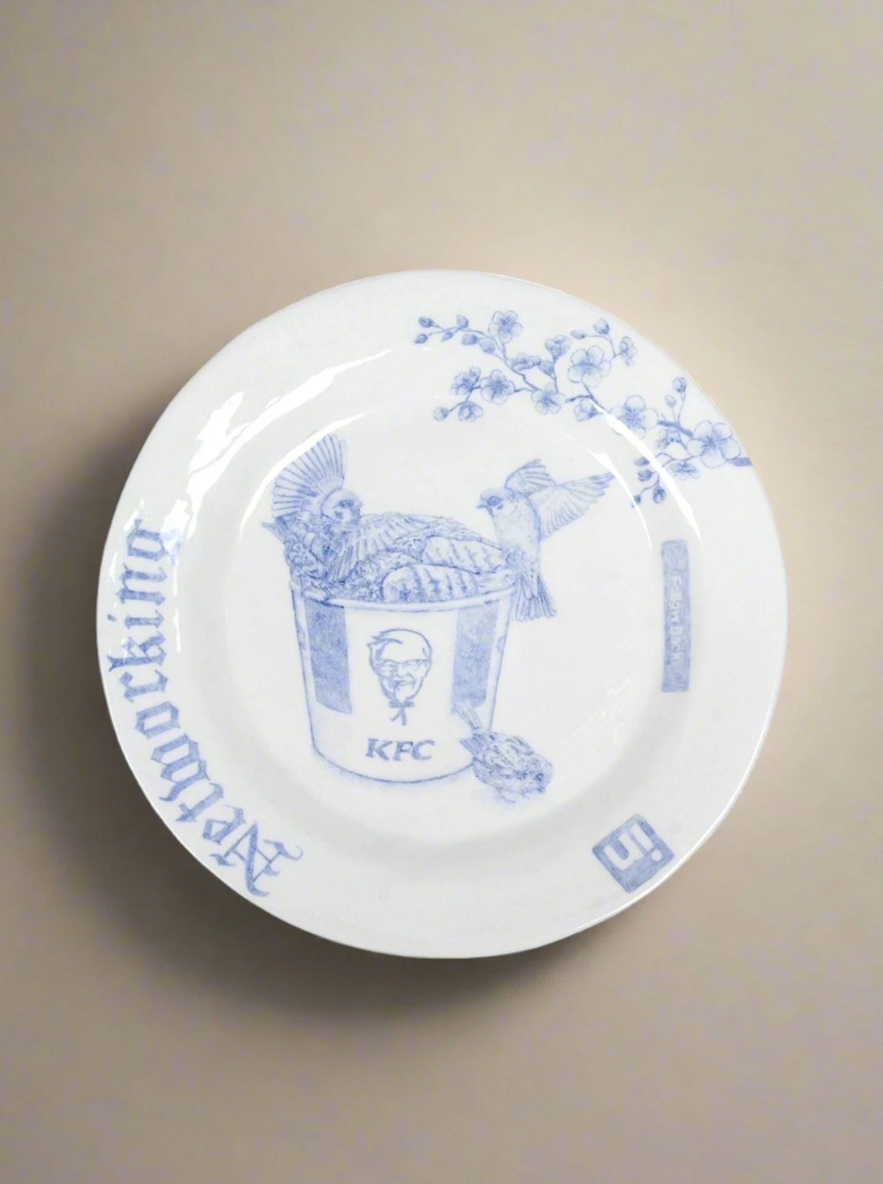 The "Networking - Decorative Dish" by Mathieu Frossard is a white porcelain plate featuring blue underglaze designs that depict a KFC bucket filled with a chicken skeleton. A bird perches atop the bucket, while branches adorned with leaves and berries extend above. The intricate hand-drawn illustrations are complemented by Japanese characters and the number "11.