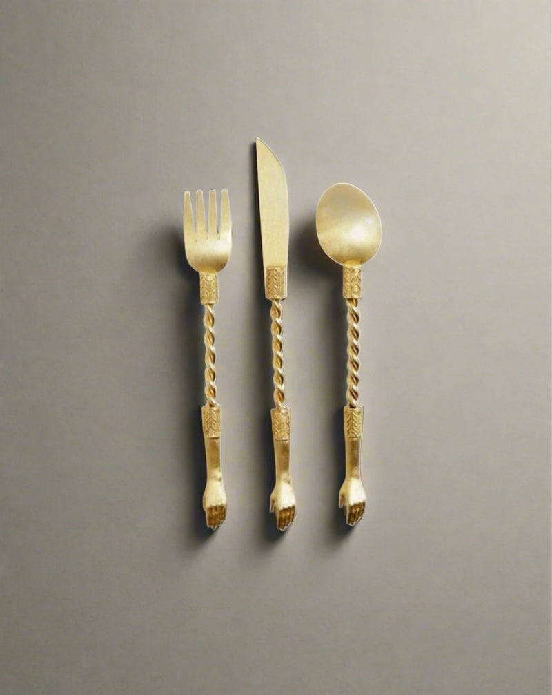 The Hand Set by Natalia Criado includes a gold-colored fork, knife, and spoon with elegantly twisted handles. This handcrafted collection enhances any dining experience with luxurious sophistication, set against a plain grey background.