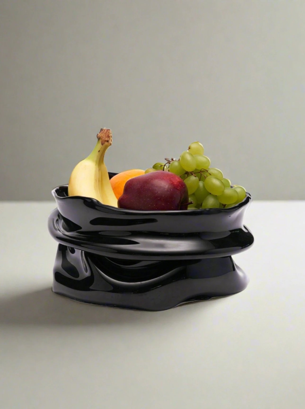 The Nina Fruit Bowl in Resin by Studio X, with its black, glossy sculptural design, elegantly holds a bunch of grapes, a banana, a plum, and an orange. The handcrafted piece stands out against the neutral gray wall and light-colored surface.