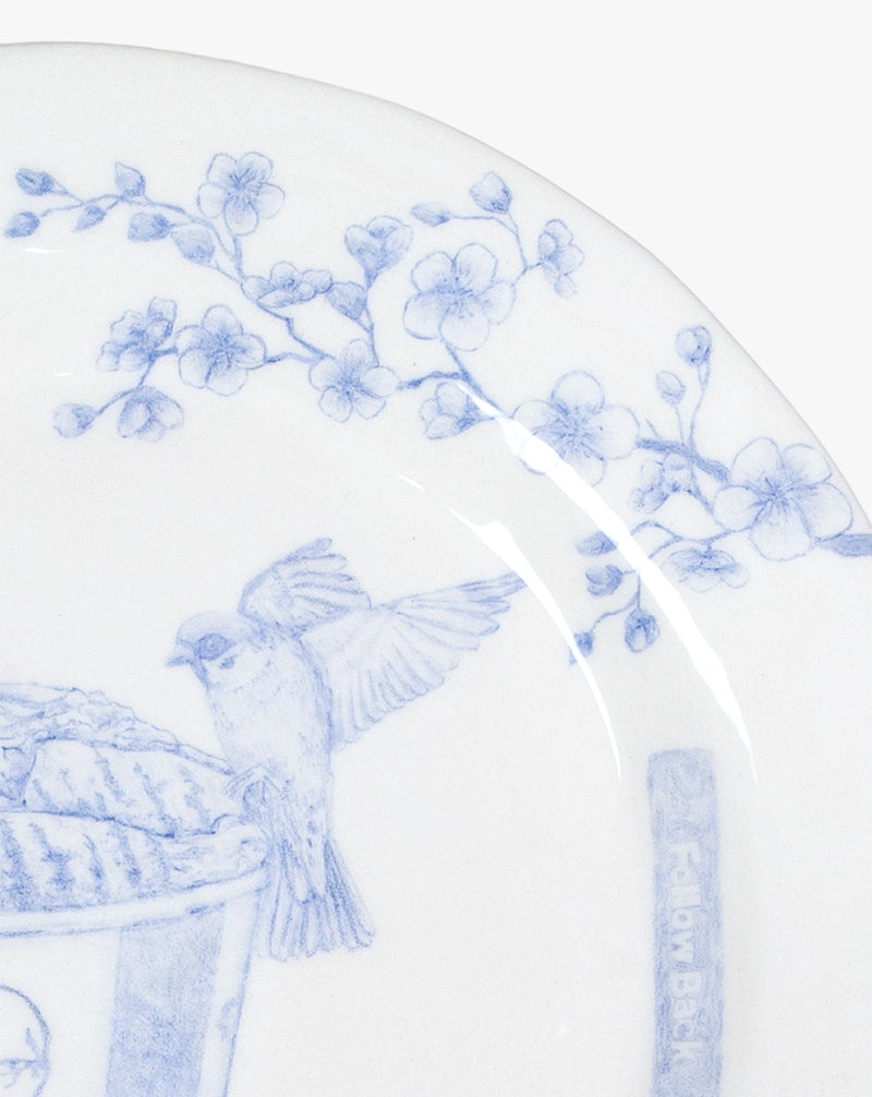 A close-up of the Networking - Decorative Dish by Mathieu Frossard featuring a blue and white design. The design includes a bird perched on the edge of a cylindrical container, with hand-drawn illustrations of cherry blossoms extending across the plate. The underglaze artwork is delicate and finely detailed.