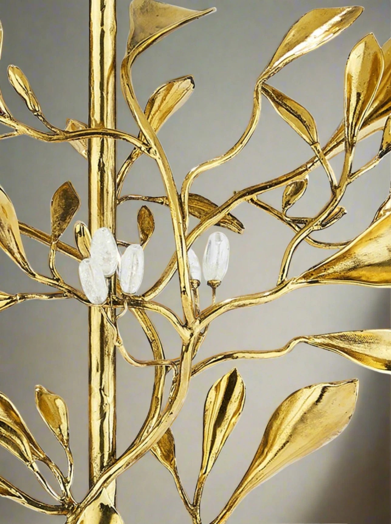 A close-up of a Foliage Table Lamp by Goossens Paris showcases a handcrafted gold metal design resembling tree branches with elongated leaves and three translucent white ovals. Set on a neutral backdrop, its minimalist aesthetic makes it an exquisite addition to any art collection.