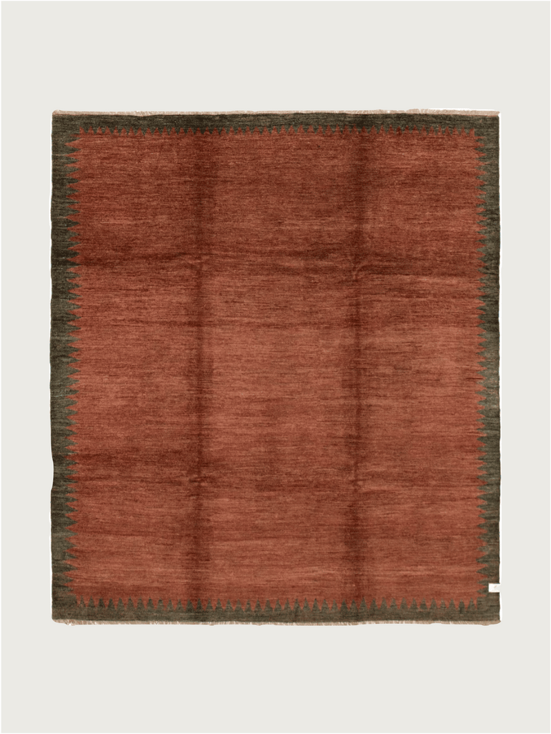 The Ziegler Rug in Carob by catalog:three is a square wool piece measuring 244 cm x 274 cm. It features a warm rust center with a dark green zigzag border, creating a hand-knotted, plush texture with subtle shading variances, set against a plain light background.