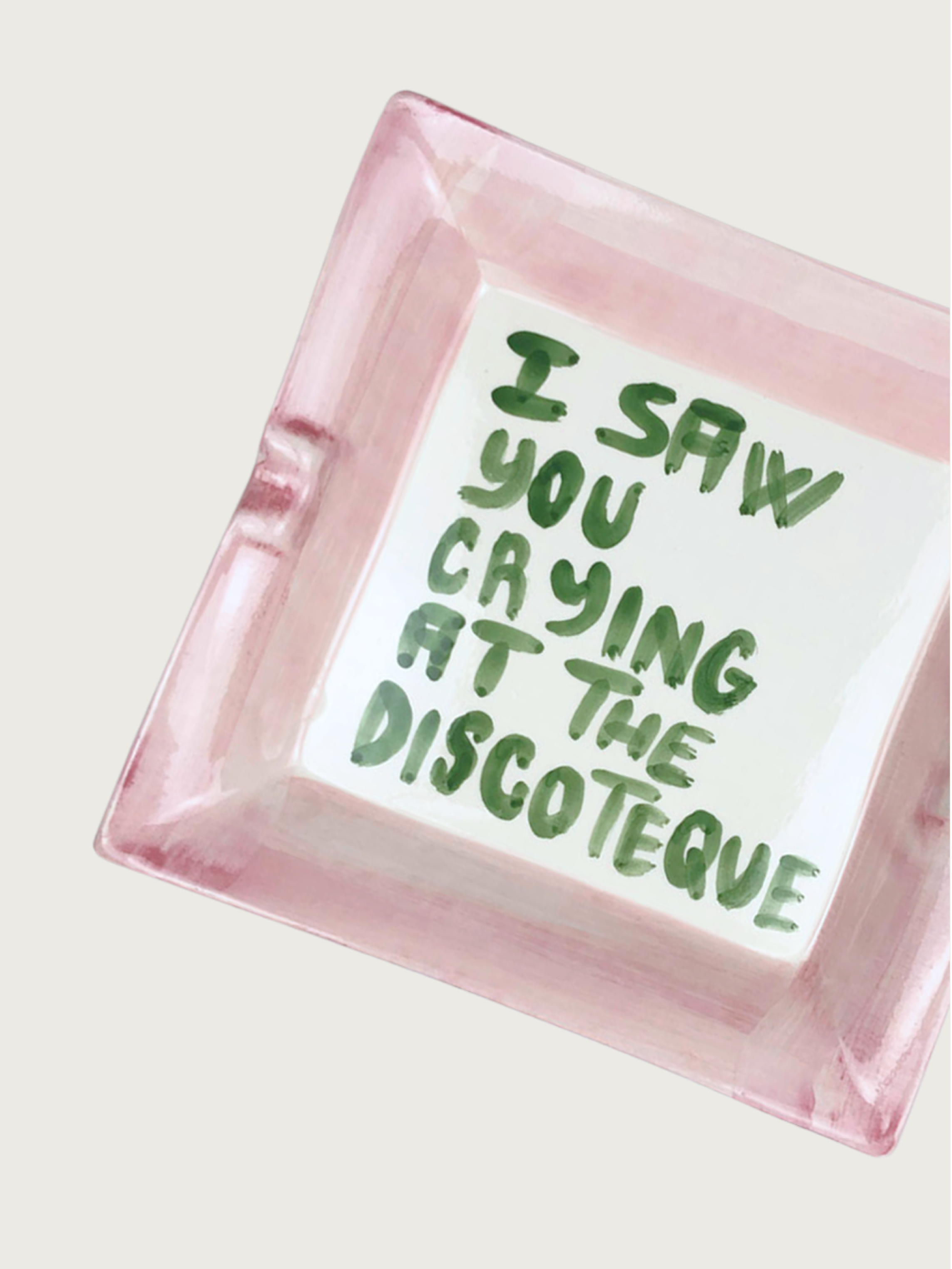 The "I saw you crying at the discoteque" ashtray by Musae Studio is square with a pink rim, featuring nostalgic, handwritten green text on a neutral beige ceramic background.