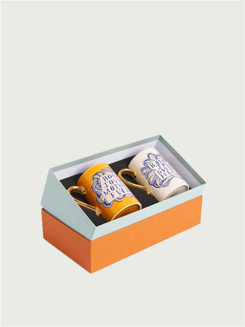The L’Objet Ruan Hoffmann Sweetheart + Mother Fucker Mugs Set of 2 comes in a gift box with an orange base and light blue lid, featuring elegant yellow and white mugs with gold handles and blue decorative text.