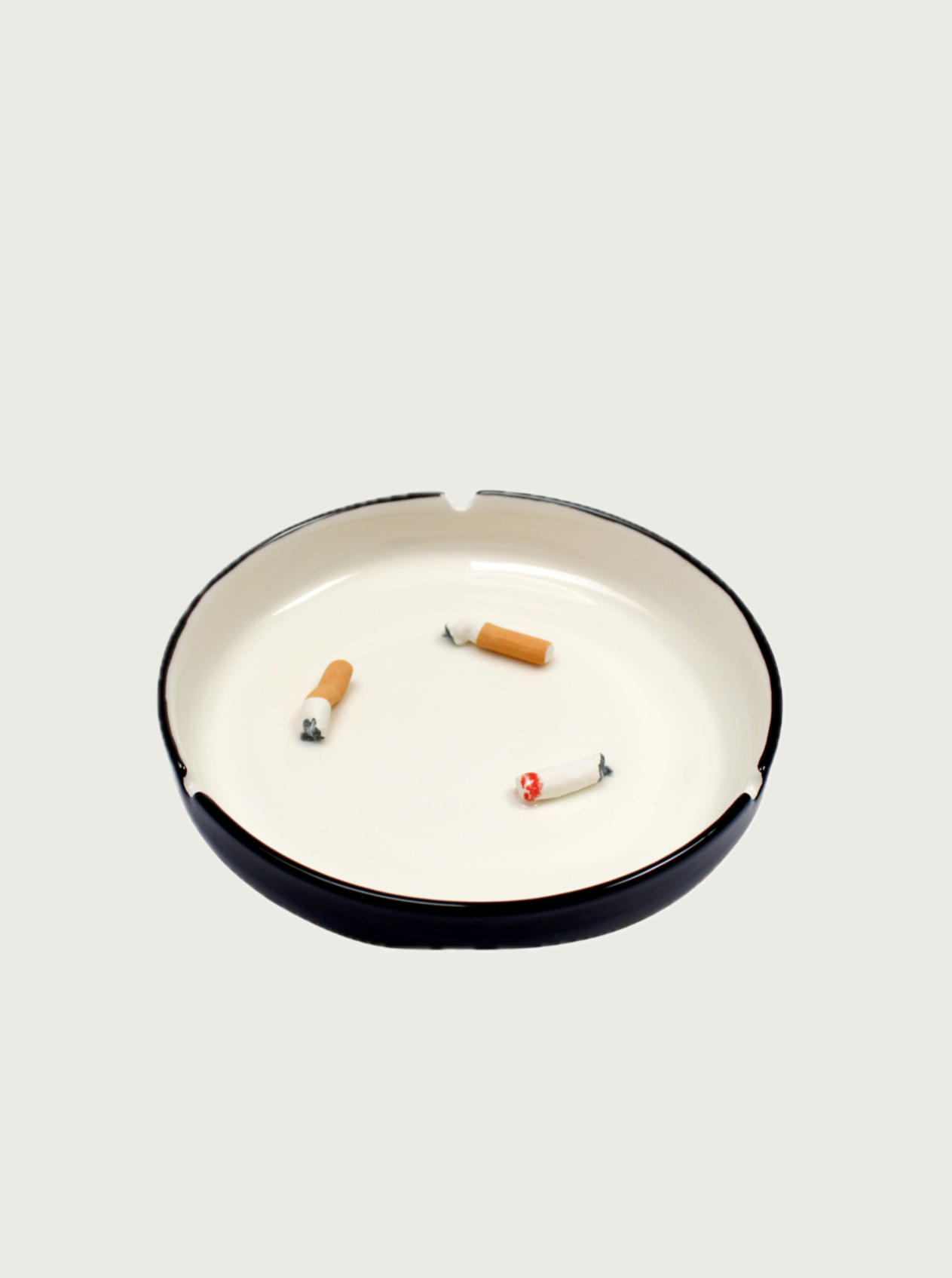 A glazed earthenware Cigarette Butts Ashtray by Villa Arev sits on a plain surface, holding four cigarette butts arranged at different angles. The background is a light cream color, reminiscent of a Mediterranean getaway at Villa Arev, and the ashtray has three notches around its rim.