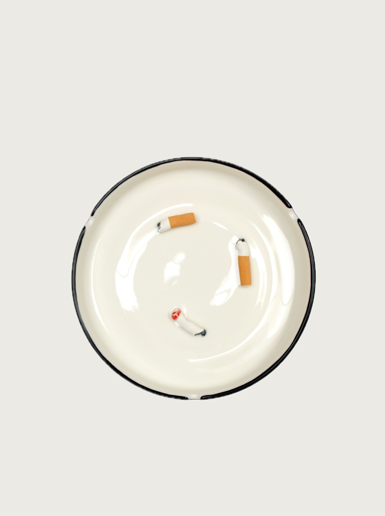 A white ceramic earthenware Villa Arev Cigarette Butts Ashtray with a black rim contains three cigarette butts, one of which is still slightly lit. The butts are scattered randomly within the ashtray on a light gray background, reminiscent of tranquil moments at Villa Arev during a Mediterranean getaway.