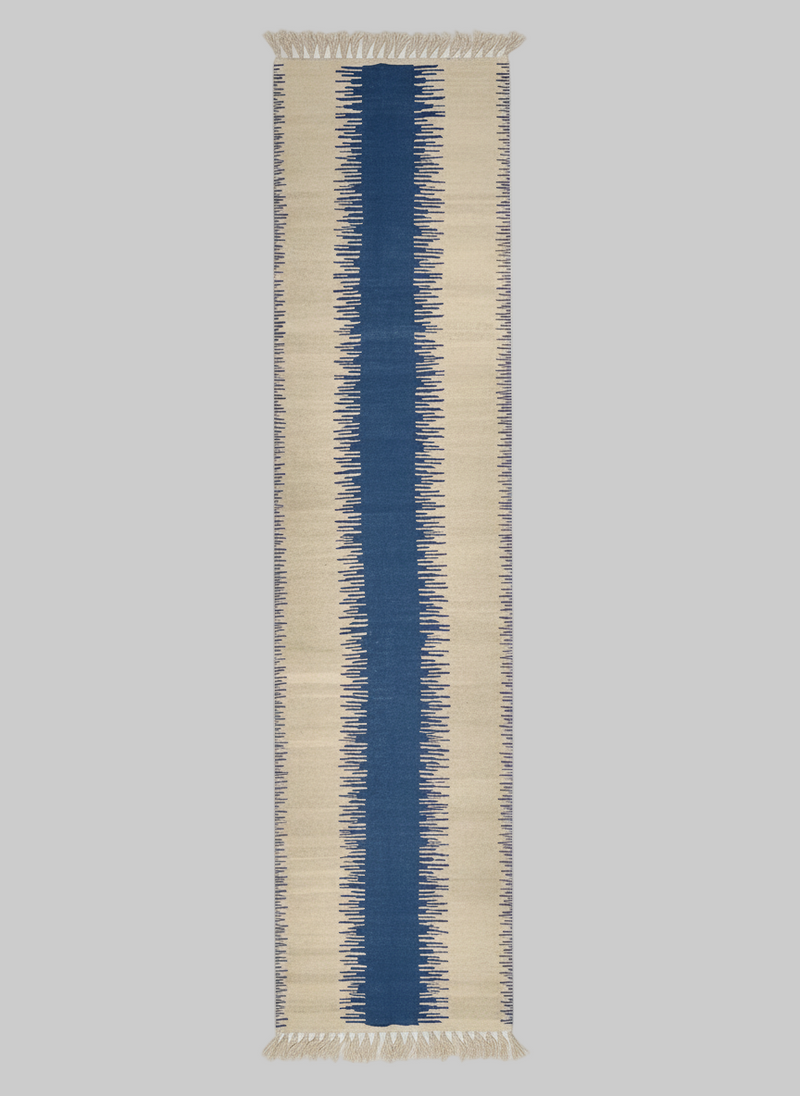 A long, rectangular Navy Anar Kilim Runner - Handwoven Rug by ISHKAR with a jagged, symmetrical pattern runs vertically down the center. This handwoven wool rug features a wide central blue stripe flanked by narrower cream-colored stripes with irregular, serrated edges, all created using natural dyes that blend into the background.