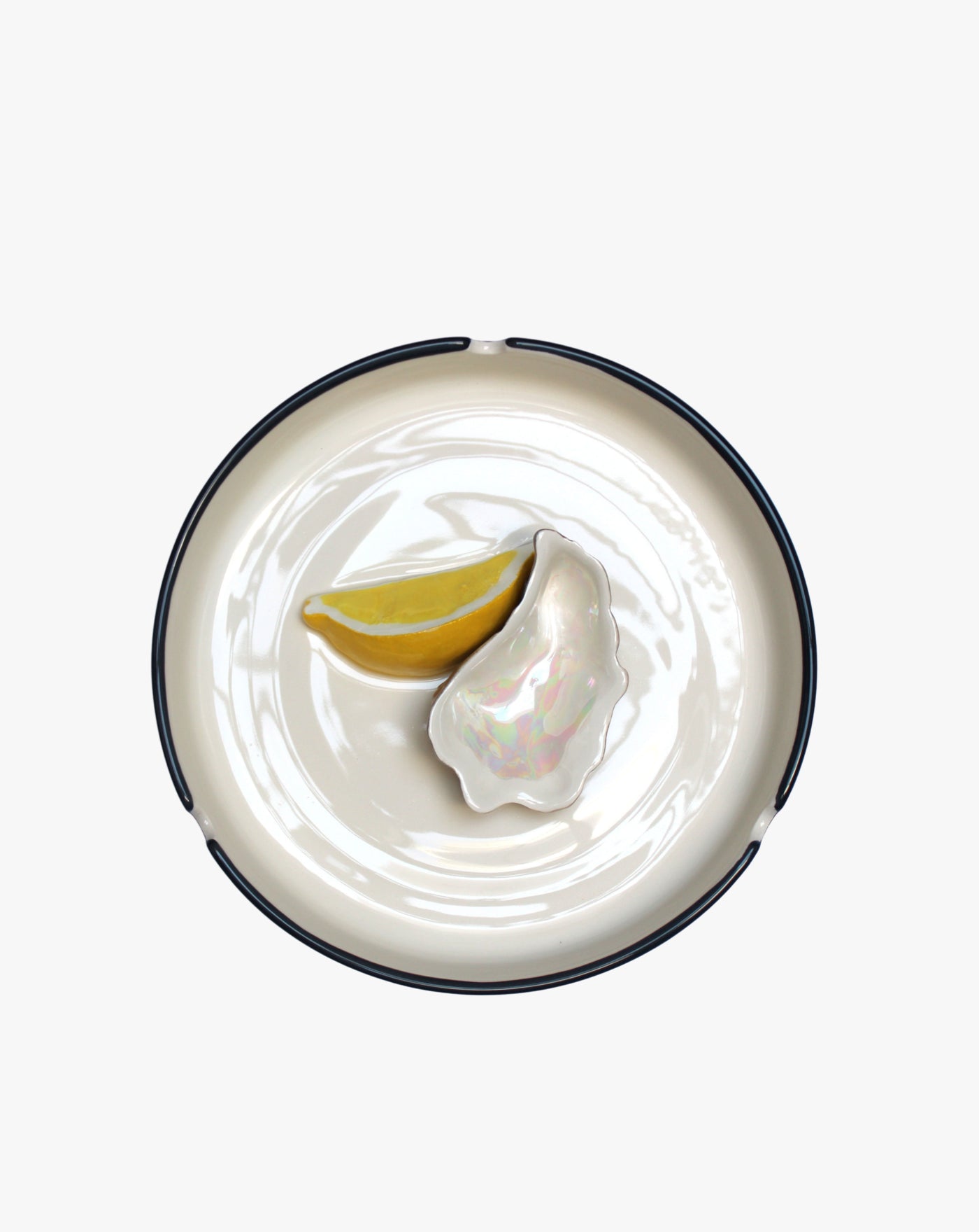 A simple white glazed earthenware Plaisir Solitaire Ashtray with a black rim, displaying a single fresh oyster accompanied by two lemon wedges on a plain background. Brand: Villa Arev.