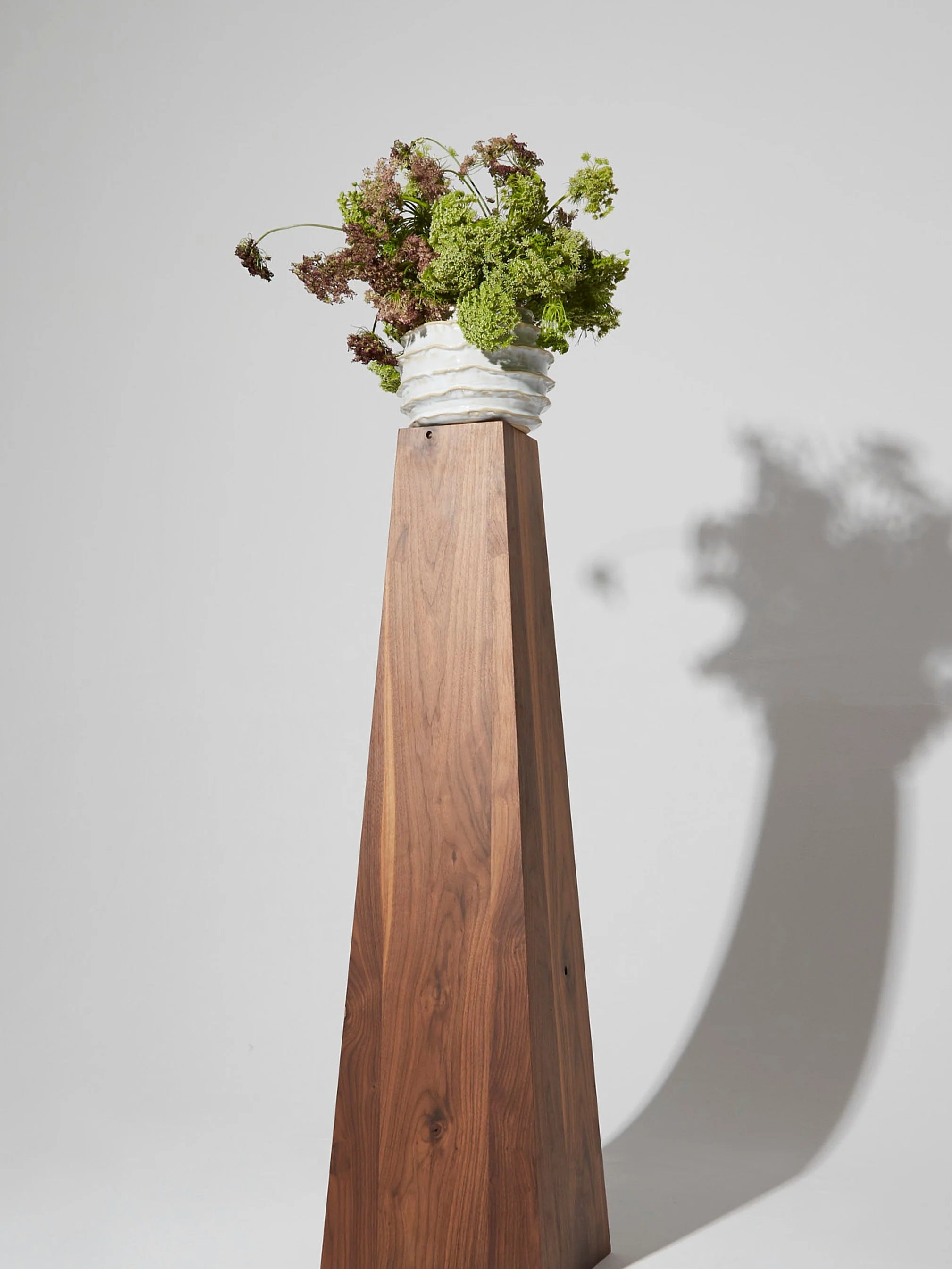 A wooden pedestal supports the Project 213A's Holiday Vase in Shiny White, which is filled with a lush arrangement of greenery. The shadow of the vase and plants is cast on the light gray background.