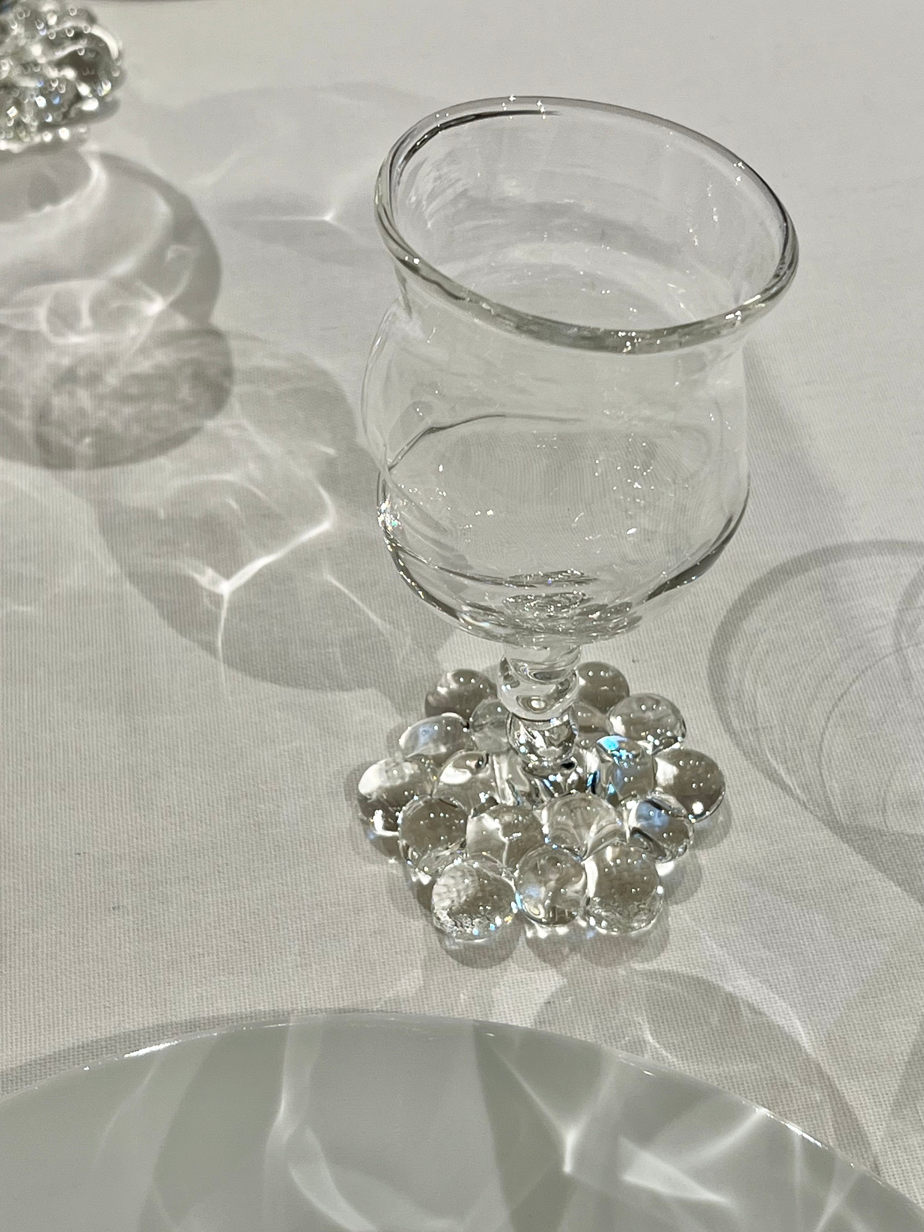 An elegant Justine Menard Mouthblown Wine Glass Set on a white tablecloth, surrounded by decorative clear and iridescent beads that sparkle under soft lighting, casting intricate shadows.