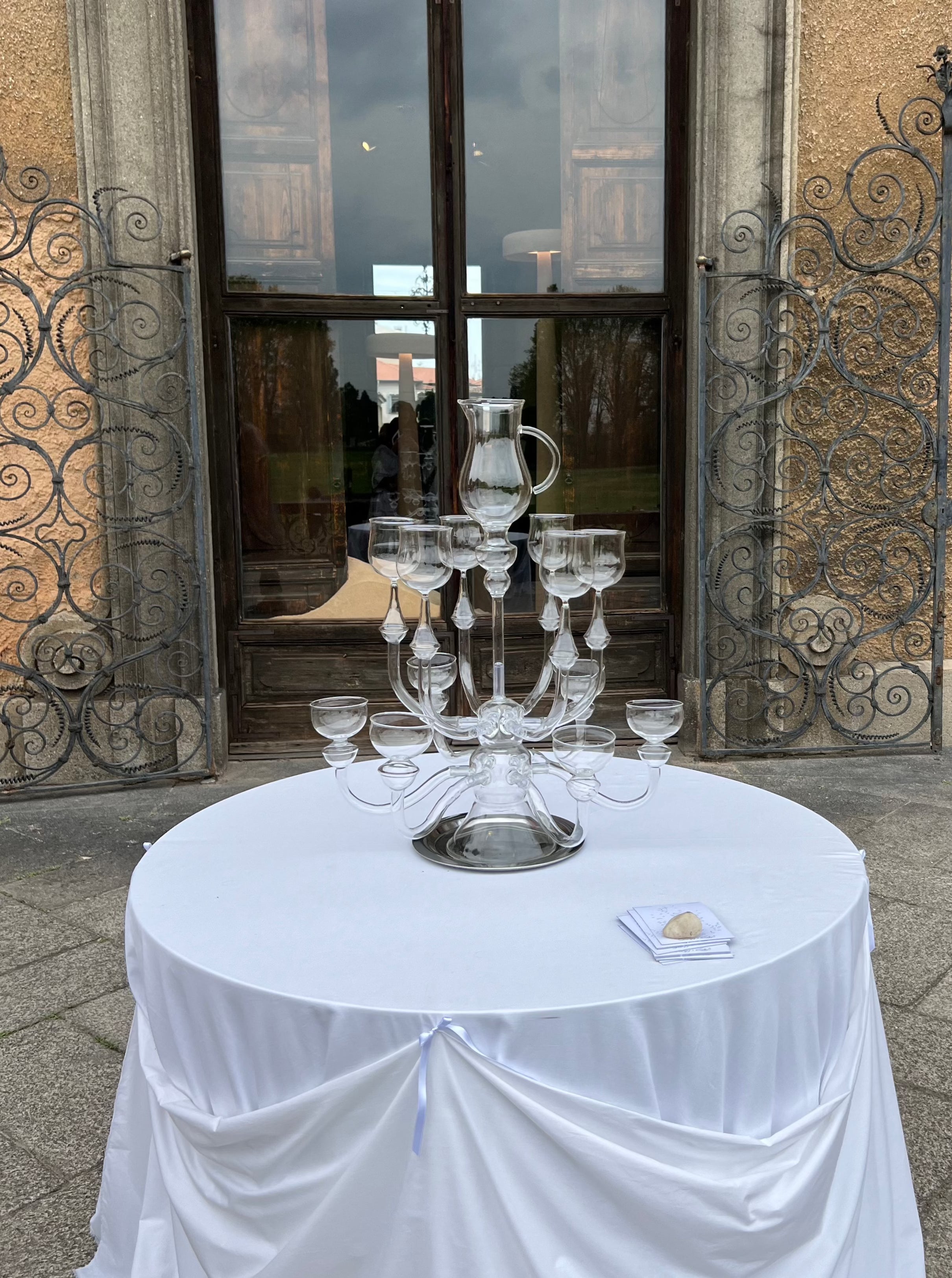 A round table covered with a white cloth stands in front of ornate metal gates. On the table is a large, tiered "Dinner's Ready" Glass Sculpture by Ylaria Pavone, surrounded by elegant glass dinnerware. A folded piece of paper and a small stone lie on the tablecloth adjacent to the centerpiece, ready for a unique dining experience.