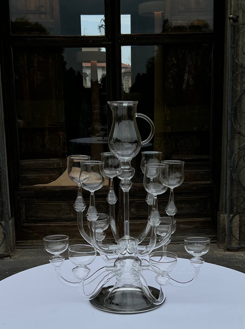 A clear glass centerpiece titled "Dinner's Ready" by Ylaria Pavone, featuring an intricate design with multiple candle-like holders arranged in tiers around a central pitcher, sits on a round white table. The background showcases a reflective glass door with dimly lit interior and exterior scenery, enhancing this unique dining experience.