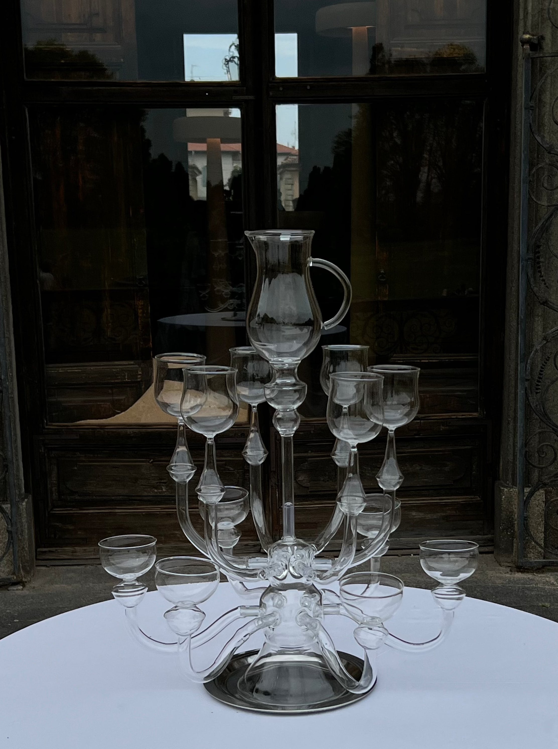 A clear glass centerpiece titled "Dinner's Ready" by Ylaria Pavone, featuring an intricate design with multiple candle-like holders arranged in tiers around a central pitcher, sits on a round white table. The background showcases a reflective glass door with dimly lit interior and exterior scenery, enhancing this unique dining experience.