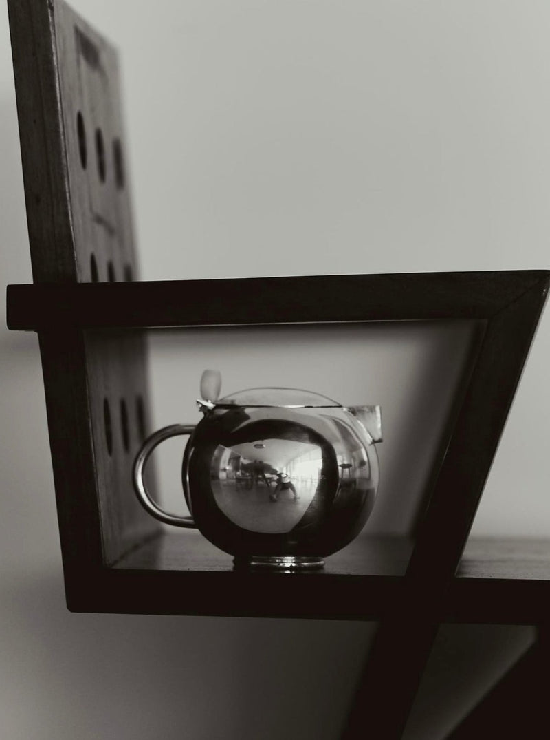 A monochrome image showing a clear, round Natalia Criado Teiera on a wooden shelf. The teapot reflects an inverted image of its surroundings during teatime gatherings.