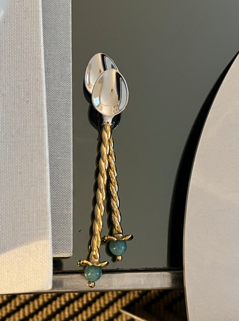 A pair of Venise Dessert Spoons from LOBJET, featuring twisted gold handles and decorative blue beads, rests on a glossy surface. A light-toned fabric with ornate patterns partially peeks in for added sophistication.