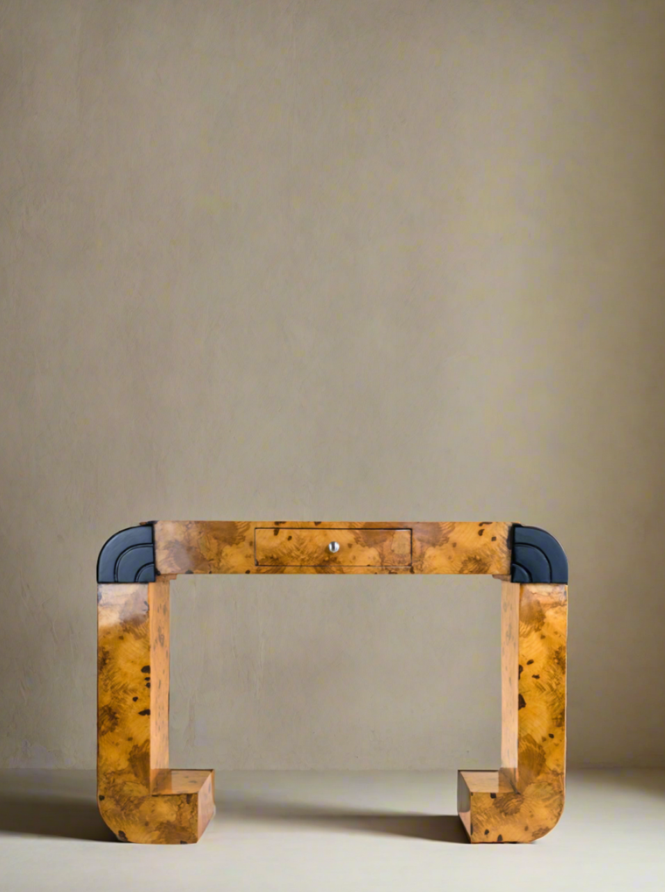 A modern wooden desk with an arch-like design, featuring one central drawer with a small knob. The handmade Serif Desk Burl Wood by Meola Interiors has a natural wood finish and curved sections at its base, complemented by dark blue accents on the sides. This piece channels mid-century design and is placed against a plain beige wall.