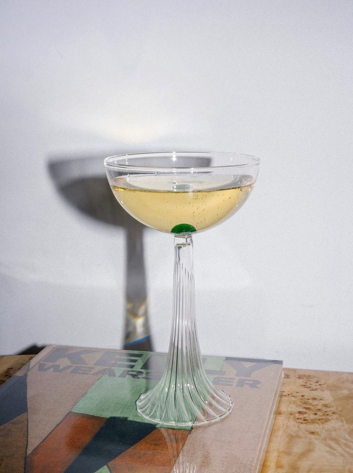 A martini with an olive sits atop a handmade Agustina Bottoni borosilicate glass stem, placed on a book with a colorful cover. The Nereida — Coupe Glass casts a soft shadow on a light background.