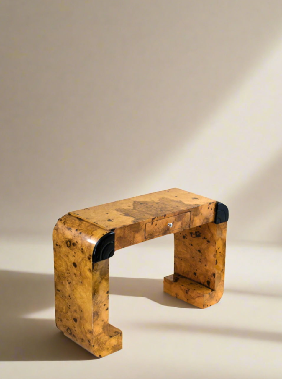 A small, minimalist wooden desk with a unique, irregular burl wood grain pattern. Its curved, arch-like structure and mid-century design have no visible drawers or compartments. Placed against a beige background with soft light creating subtle shadows, the Meola Interiors Serif Desk Burl Wood epitomizes handmade craftsmanship.