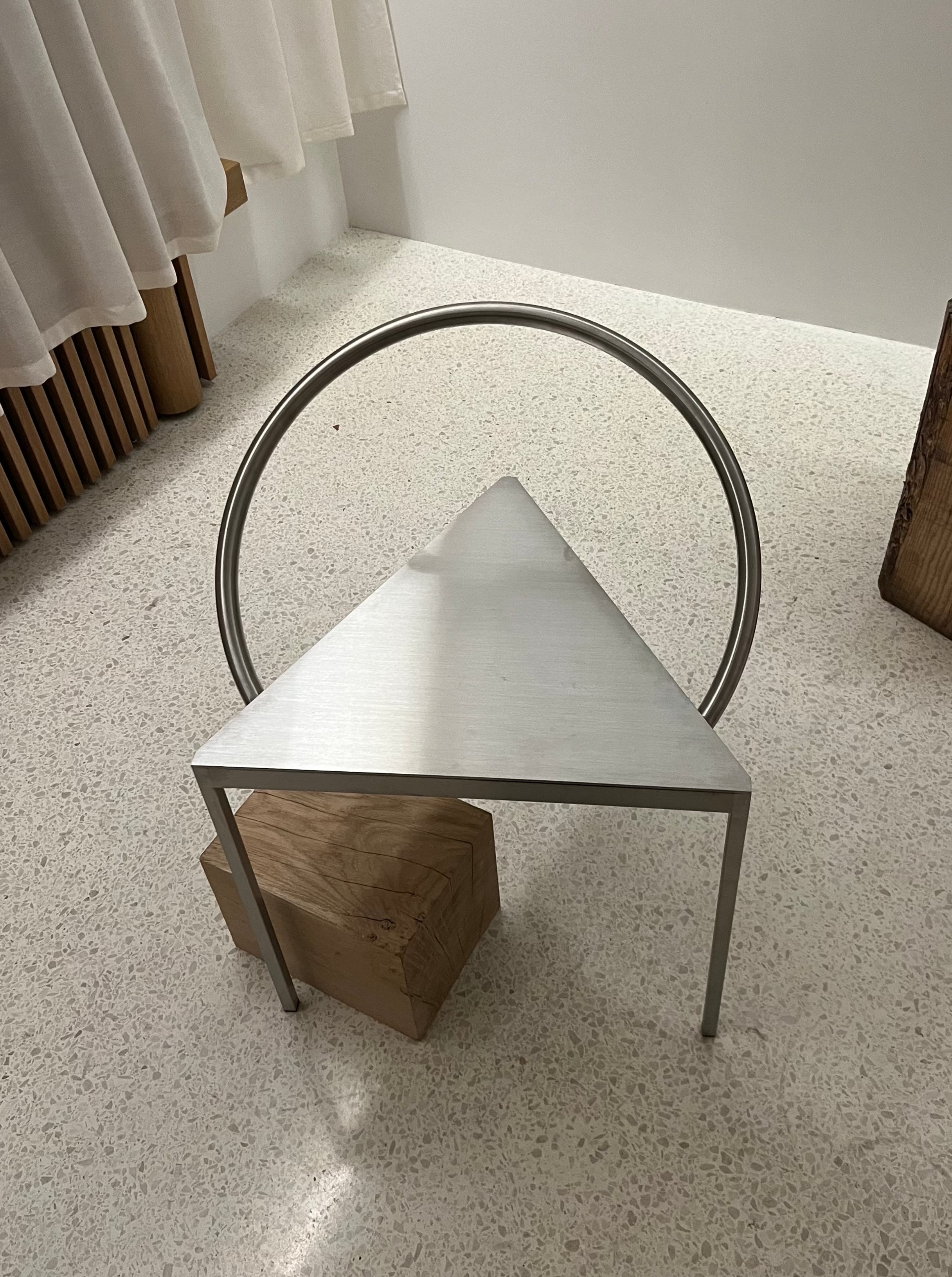 A modern art sculpture featuring a Triangolo Chair | Stainless Steel by FRAMA with a triangular metallic shape supported by two legs, resting on a wooden block. Behind the triangle is a circular metal ring, and the whole setup exudes an industrial aesthetic. The sculpture is placed on a speckled floor, with curtains and a wooden piece in the background.