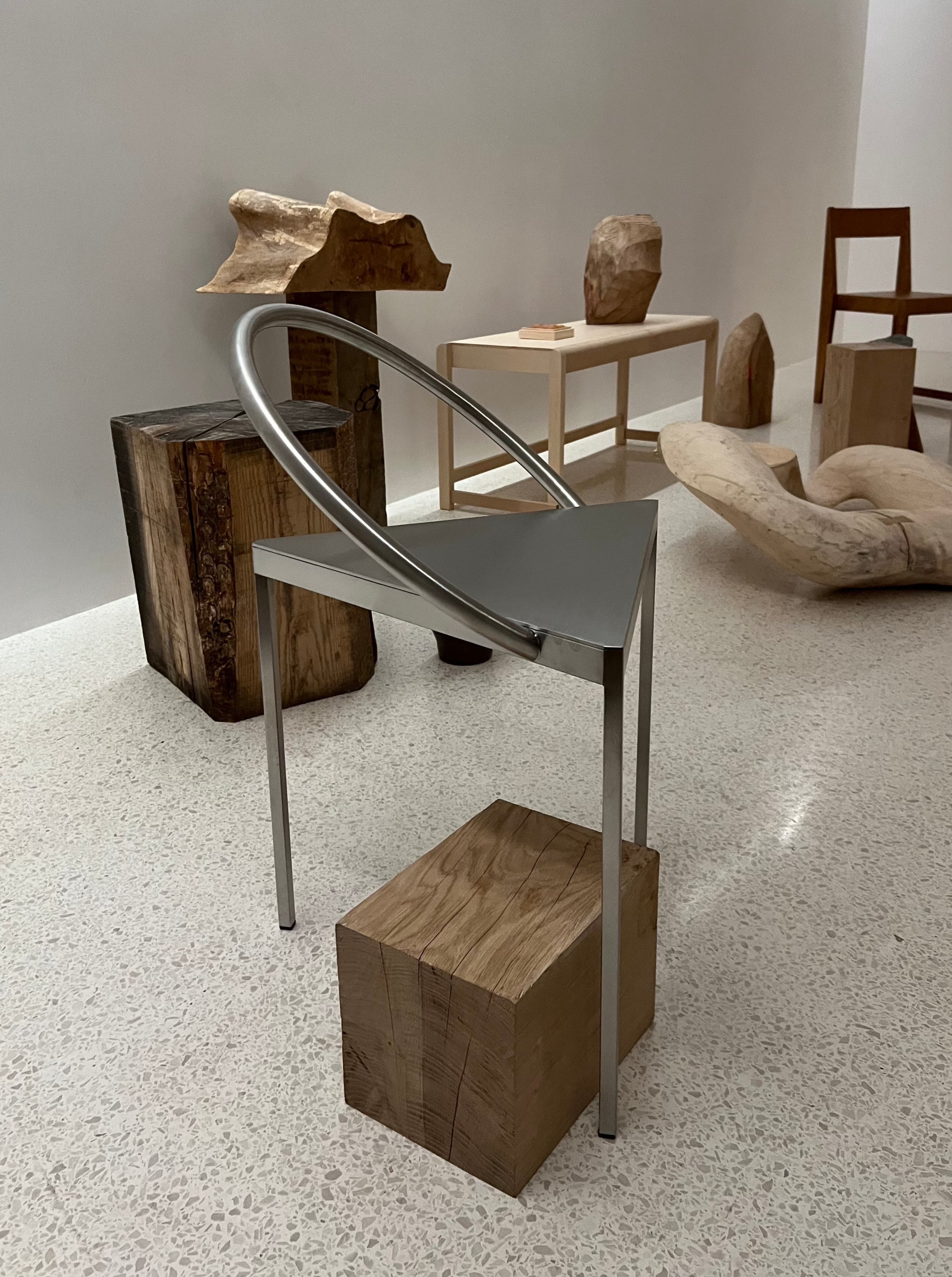 A modern art exhibit featuring the Triangolo Chair | Stainless Steel by FRAMA, with a unique aluminum loop for the backrest, set atop a wooden block. Surrounding the chair are various wooden sculptures and minimalist furniture pieces, all echoing an industrial aesthetic in a well-lit gallery space with a speckled floor.