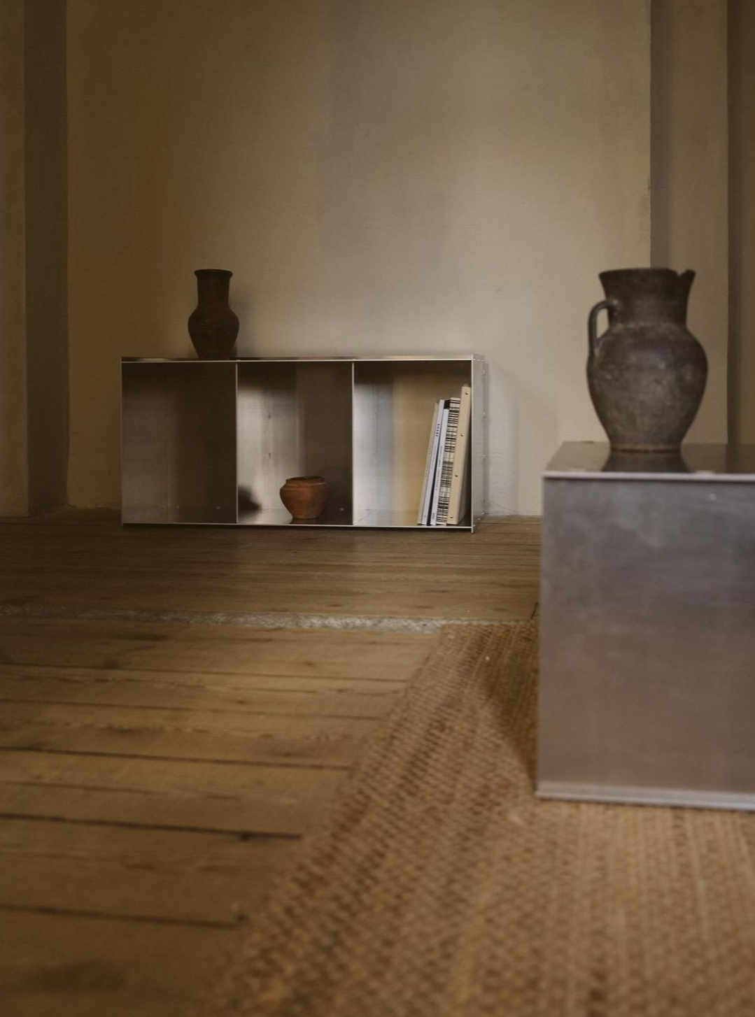 A minimalist room with wooden flooring and a woven rug. It features a low metal bookshelf with an assortment of vases, handcrafted aluminum accents like the Rivet Case | Aluminum from FRAMA, and a couple of books. The wall behind is plain, adding to the versatile design of the space. The Instagram interface is visible.