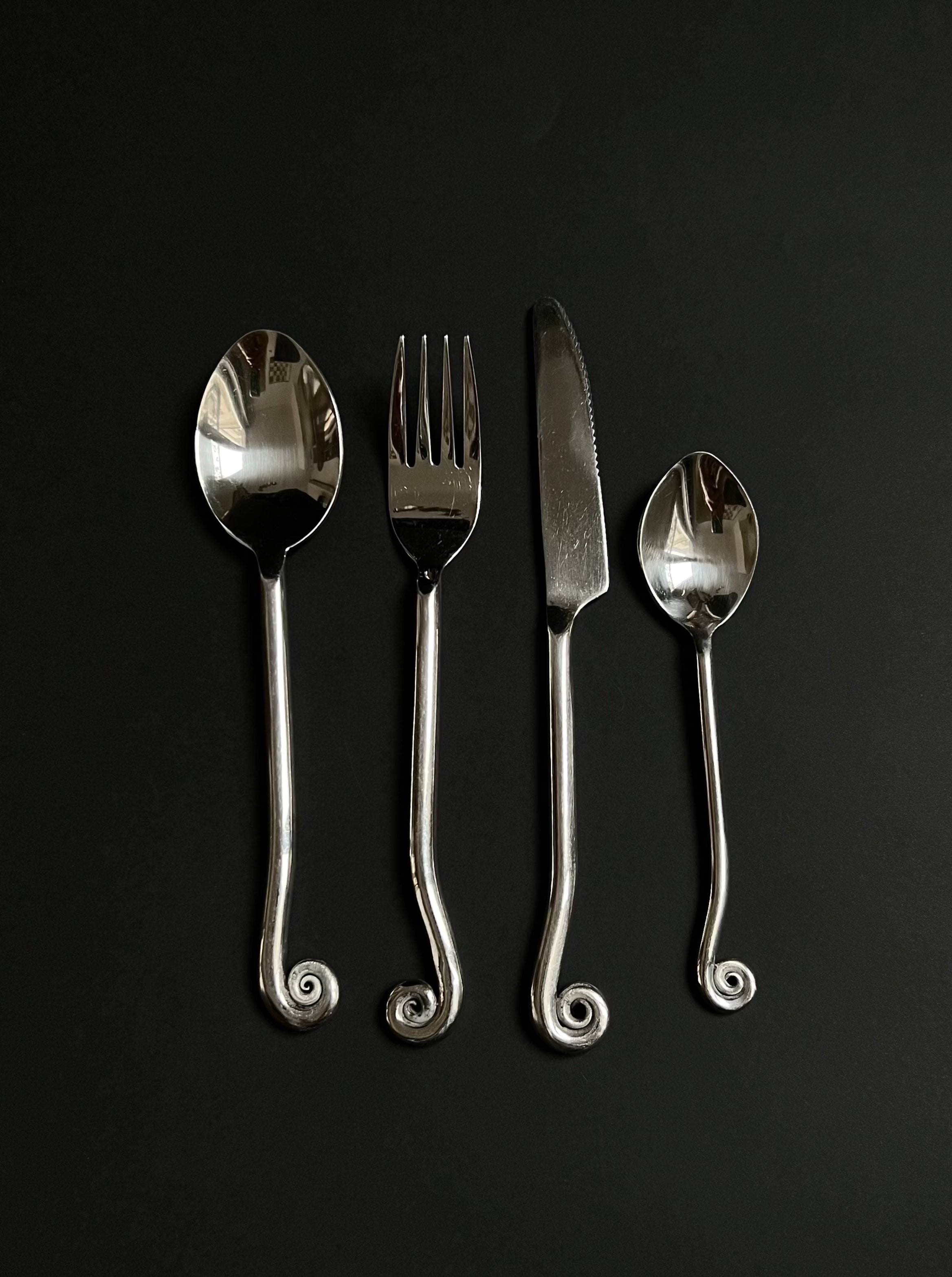 Four pieces of Manushi's Vintage Swirl Cutlery set—a spoon, fork, knife, and another spoon—with timeless design and spiral handles arranged neatly on a dark, matte surface.