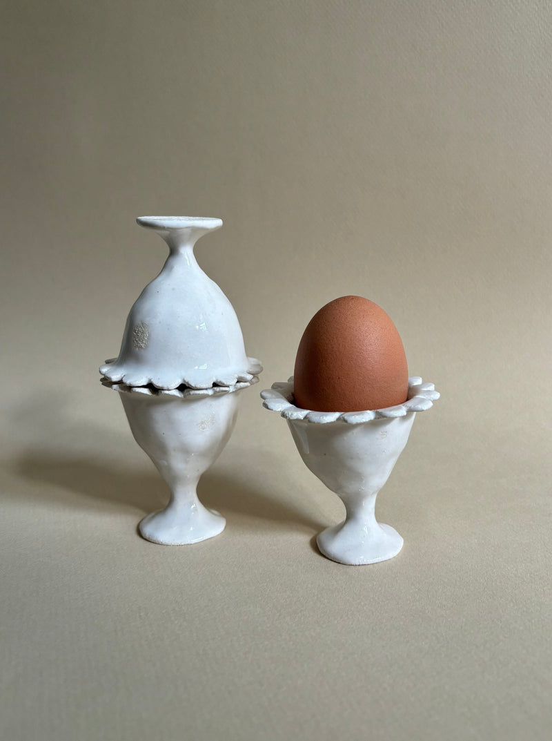A brown egg is placed in a handbuilt "Rosetta" Egg Cup by Studio Beurre, made of white clay with a decorative edge, while another matching upside-down ceramic egg cup, covered in shiny white glaze, sits beside it. The background is a smooth, warm-toned surface that complements the ceramic pieces.