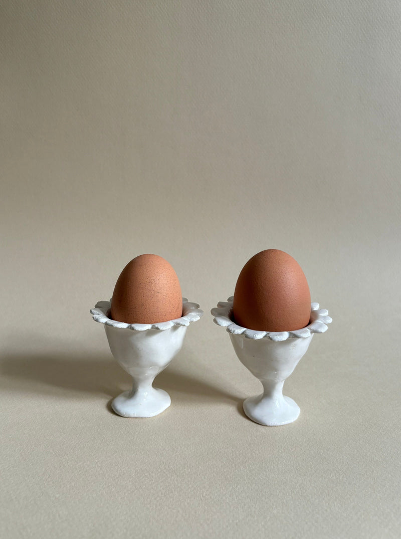 Two brown eggs are placed in white, ornate egg cups. The "Rosetta" Egg Cups, crafted by Studio Beurre from white clay with a shiny white glaze, add an artisanal touch. The background is a plain light beige, providing a simple and neutral setting that emphasizes the eggs and their holders. Soft lighting creates gentle shadows.
