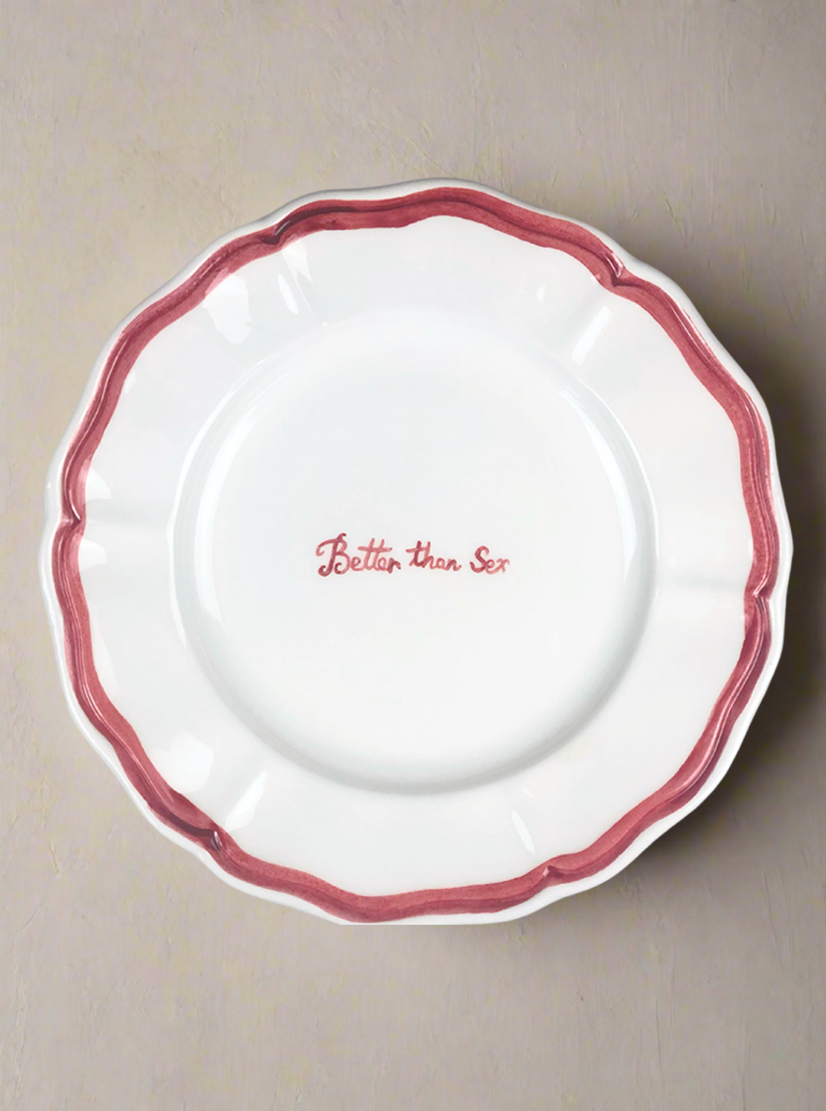 The "Better than Sex" Fil Rouge Plate by Musae Studio is a hand-painted decorative white plate with a wavy red rim and the playful phrase "Better than Sex" in elegant red cursive at its center, perfect for adding whimsy to any space.