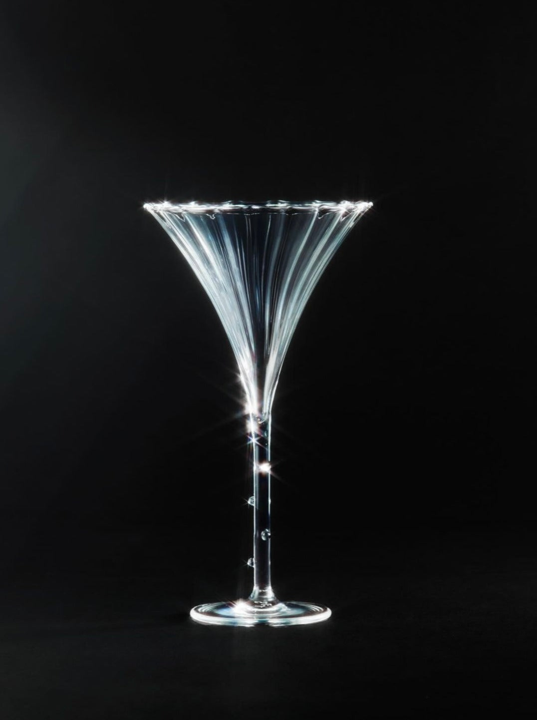 A sleek, tall Trumpet Aperitif Glass - Set of Four by Sophie Lou Jacobsen stands against a black background, catching light and creating a shimmering effect. The glass has a narrow stem and flares out at the top, resembling a classic cocktail or martini glass. Perfect for your set of 4, it adds elegance to any occasion.