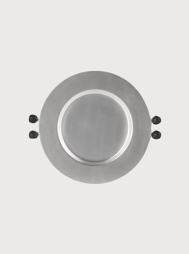 A top view of a round stainless steel Natalia Criado Lava Plate with two black handles on opposite sides. The background is a plain off-white color.