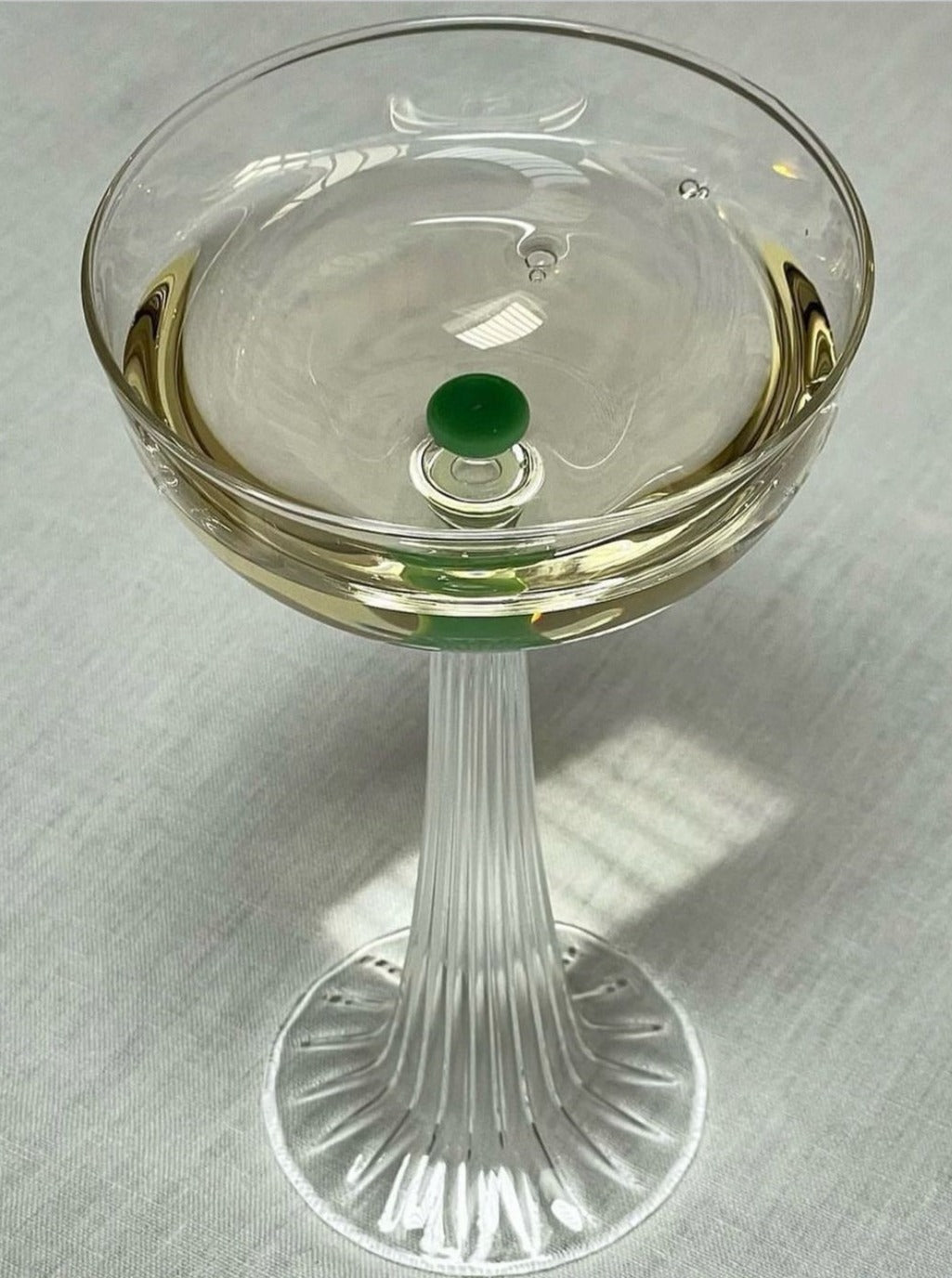 A Nereida — Coupe Glass by Agustina Bottoni, handmade in Italy from borosilicate glass, sits elegantly on a light-colored tablecloth. The clear glass, adorned with a small green garnish and etched with vertical lines on its stem and base, casts a delicate shadow as it holds either a cocktail or wine.