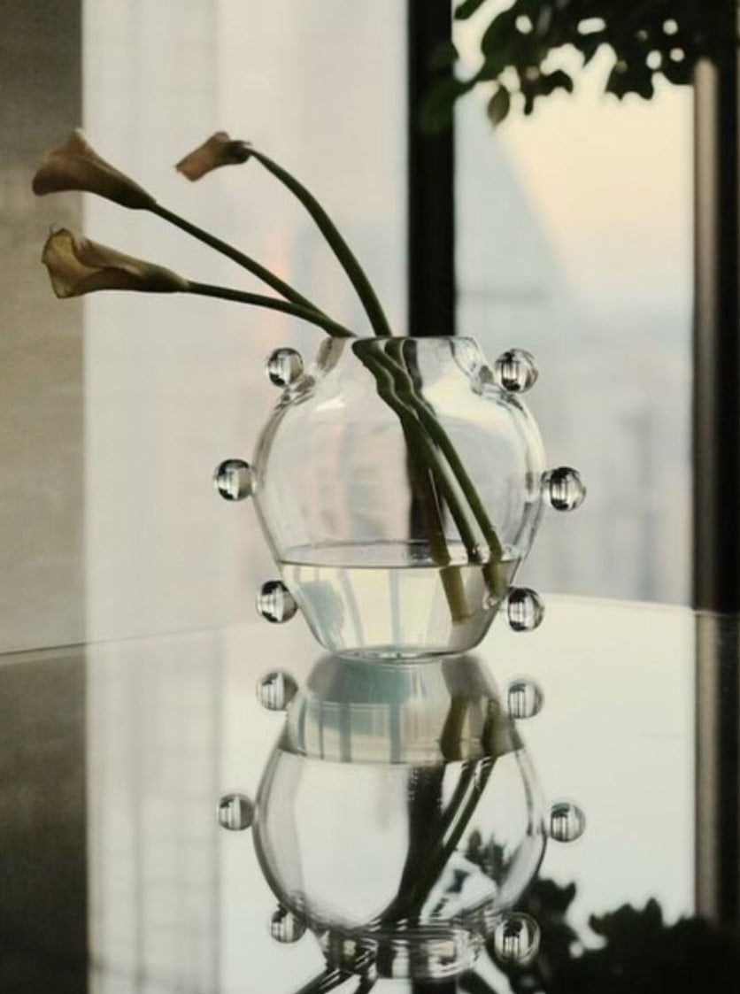 A round, clear glass hand blown La Lumiere Vase by Sophie Lou Jacobsen with small spherical protrusions sits on a reflective surface, holding three slightly drooping calla lilies. In the background, tall windows allow daylight to softly filter in, with a partial view of a tree and the outside landscape. This unique work of art showcases craftsmanship using the highest quality materials.