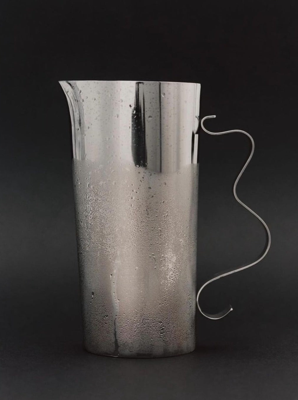 A tall, sleek stainless steel Squiggle Pitcher by Sophie Lou Jacobsen with a unique, curvy handle is shown against a dark background. Condensation droplets are visible on the pitcher’s surface, indicating it is cold. The design is simple and modern, making it an elegant counterpart to any Squiggle Tray or decorative vase.