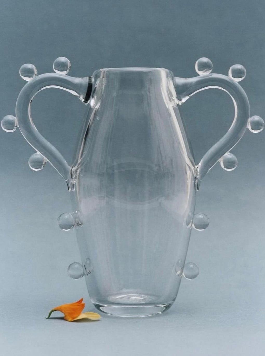 A clear, hand blown in NYC, double handled jug with two curved handles, each adorned with three small glass orbs. The body of the vase also features additional orbs arranged symmetrically. A small orange flower with green leaves lies at the base of the vase on a pale blue background. Introducing the **La Vitalité Vase** by **Sophie Lou Jacobsen**.