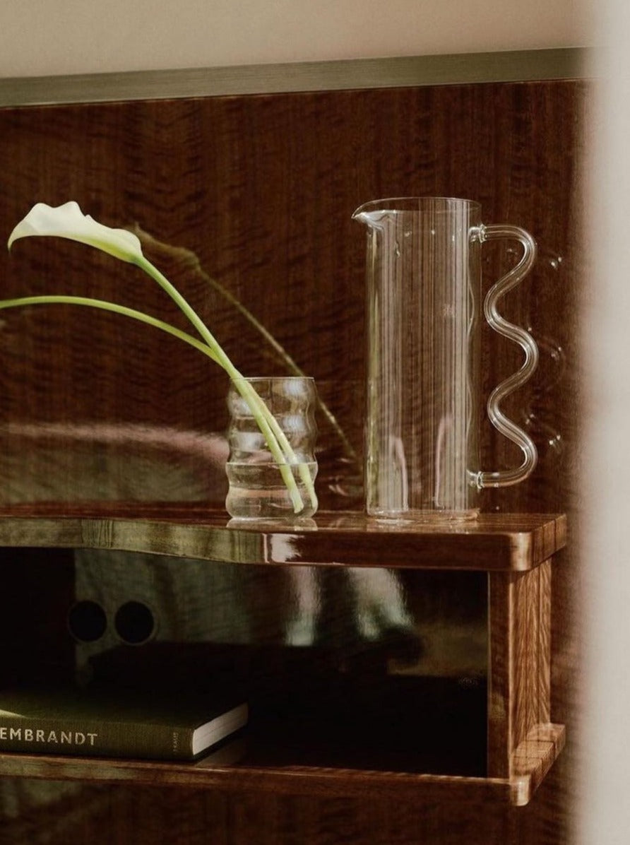 A small wooden shelf holds a transparent Wave Pitcher Glass - 9 Colours by Sophie Lou Jacobsen made of borosilicate glass, a glass holding two white calla lilies, and a book titled "Rembrandt." The shelf is against a polished wooden wall, creating a cozy and elegant ambiance.