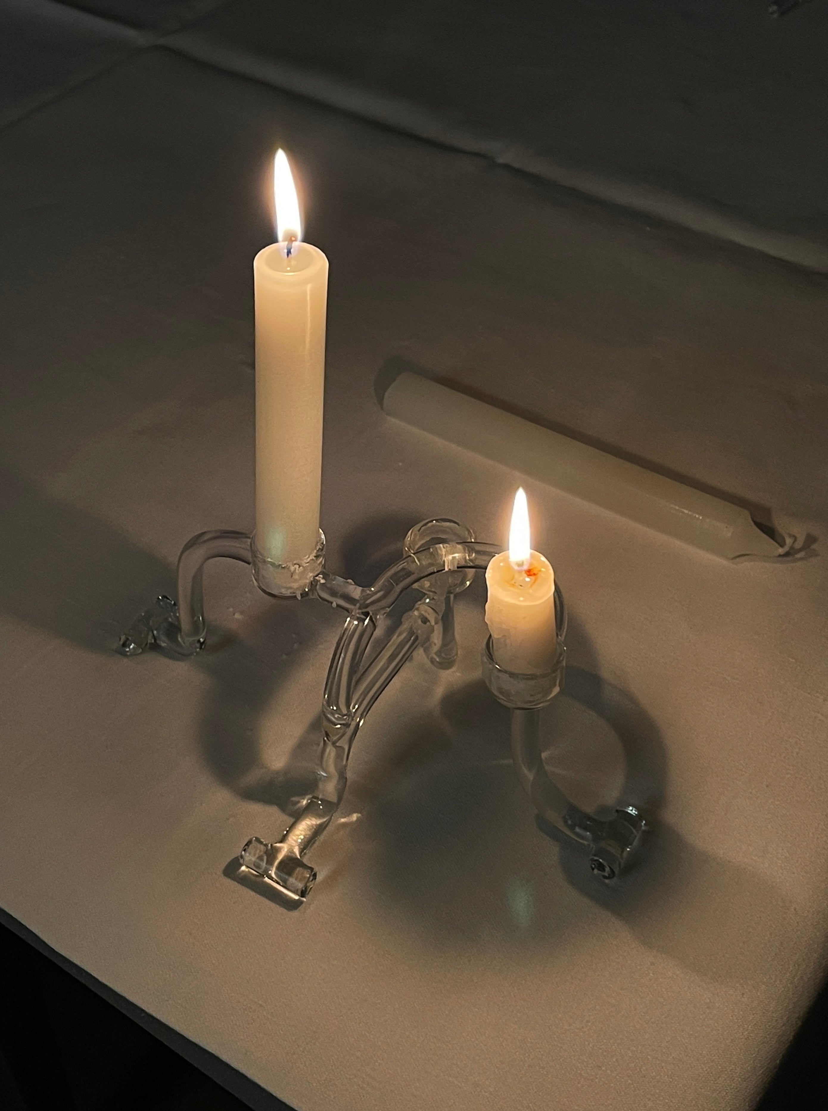 A lit candle and a shorter, melted one are held by an ornate StudioNotte Candleholders Sio2 on a dark surface. The warm glow from the flames creates a subtle ambiance, contrasting with the surrounding darkness. An unlit candle lies in the background, adding to the serene atmosphere.