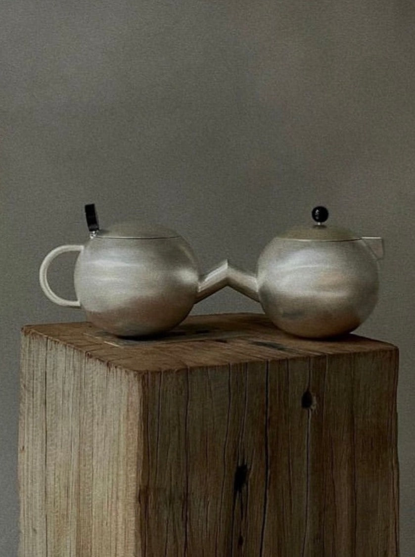 Two interconnected silver Mucura by Natalia Criado, resembling duality objects, rest on a weathered wooden block. One Mucura has a single handle and small spout, while the other has a tiny lid and spout. The background is gray and minimalistic.