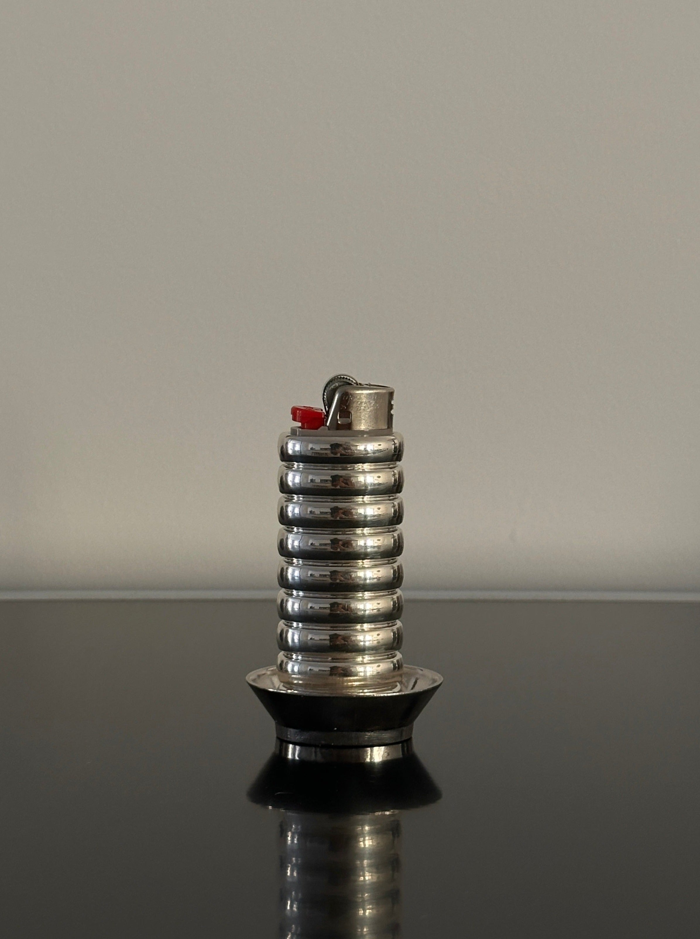 A close-up photo of a metallic, cylindrical **FRY POWERS Gio Lighter Holder** with a red button on top. The stylish lighter accessory is placed on a reflective black surface, and the background is a plain, light gray wall.