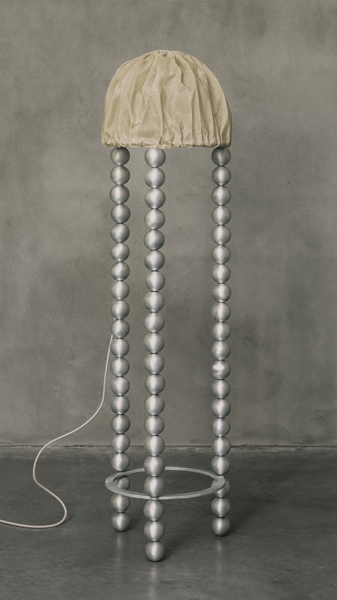 Kaytar's "ALTER" Floor Light features a spherical beaded base similar to four tall strands with subtle alchemic symbols, a round textured beige shade, and stands elegantly on smooth concrete with a visible power cord.