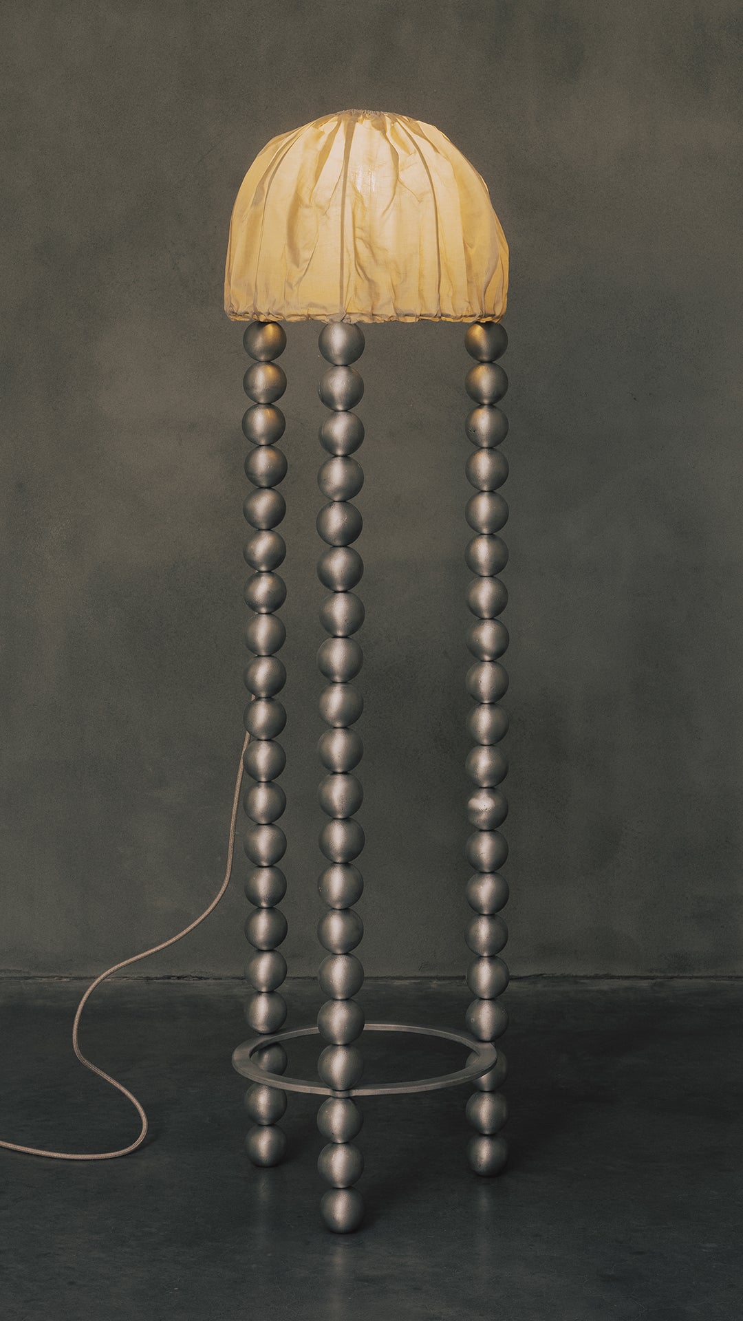 The Kaytar "ALTER" Floor Light features a tall, modern design with a dome-shaped yellow shade. Its handcrafted stand is composed of four vertical rows of metallic spheres in sand-cast metal, connected at the base by a circular ring, set against a plain dark background.