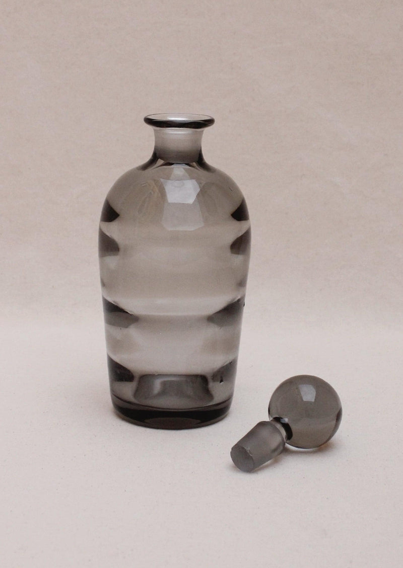 An Boga Avante Shop Art Deco translucent gray glass liquor decanter with a matching stopper, featuring a simple striped design, set against a plain light beige background.