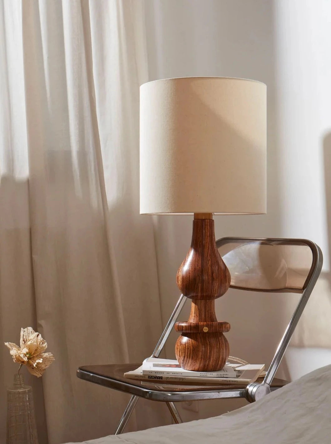 A warm, inviting scene with a Romie Objetti Lamp Marcia Zebrano turned on, casting a soft light, placed on a sleek metal framed chair. A couple of books and a dried flower accentuate the.