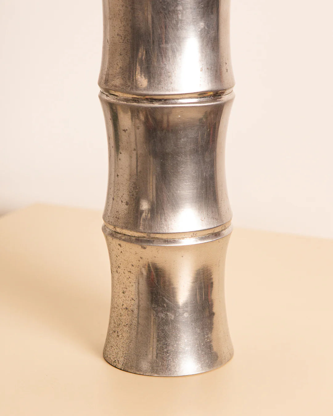 A close-up view showcases the Large Cast Aluminium Vase ‘Bambou’ 80's from Treaptyque, featuring three interconnected sections that mimic the shape of bamboo. It rests on a light-colored, shiny surface adorned with visible spots, adding a touch of vintage charm to the scene.
