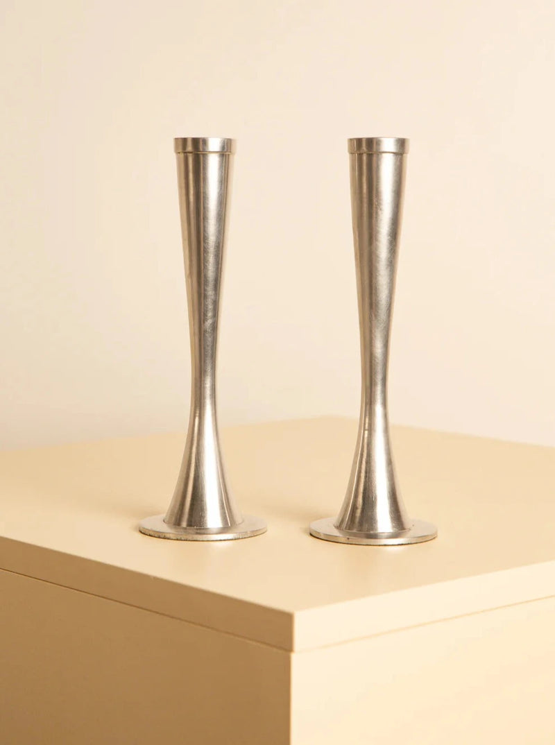 Two sleek Treaptyque Pair of 80's aluminum candleholders stand side by side on a light-colored surface. Their design is simple and elegant, with a narrow mid-section and a slightly flared base and top. The background is plain and beige, highlighting the minimalist aesthetic and showcasing meticulous craftsmanship.