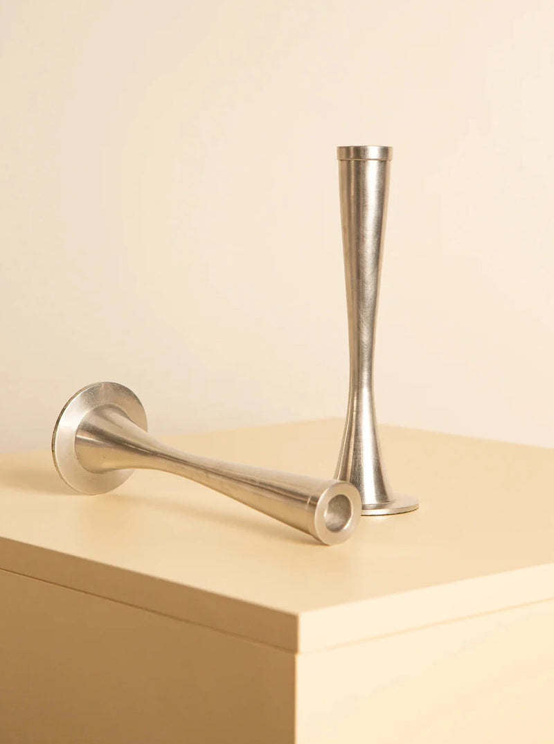 Two elegant Pair of 80's aluminum candleholders by Treaptyque are displayed on a light beige surface. One candle holder stands upright while the other lies horizontally. Their sleek, minimalist design and reflective finish, a result of meticulous craftsmanship, create a modern, sophisticated aesthetic.