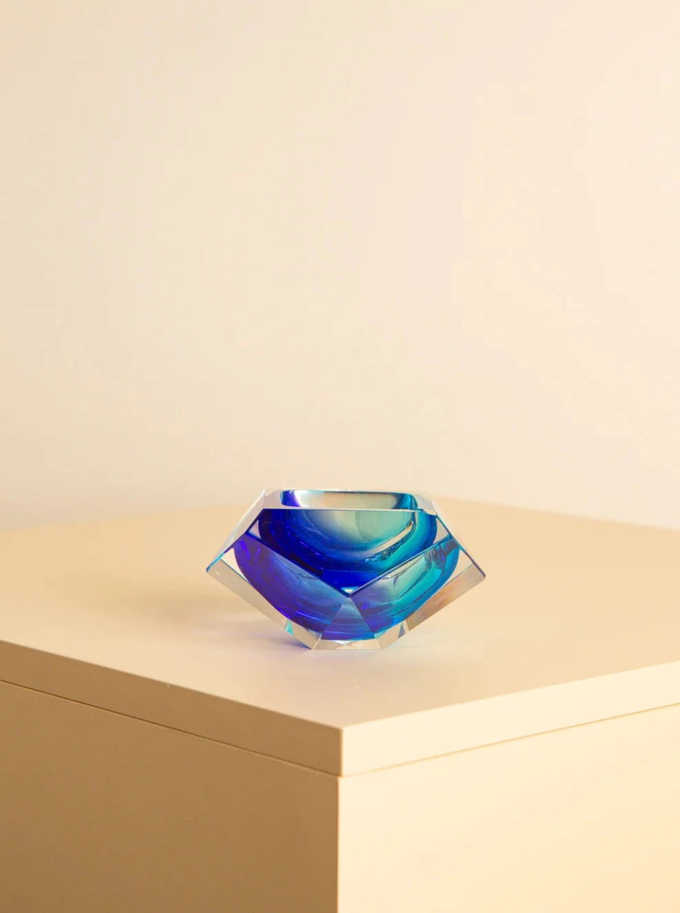 A geometric, blue-hued glass sculpture with intricate reflections and facets is placed on a light-colored, minimalist wooden surface against a plain background. The piece, reminiscent of Flavio Poli's Italian glass art, appears to be a Blue “Diamant” ashtray in murano glass by Flavio Poli for Seguso 60's. This exquisite item is offered under the Treaptyque brand name.