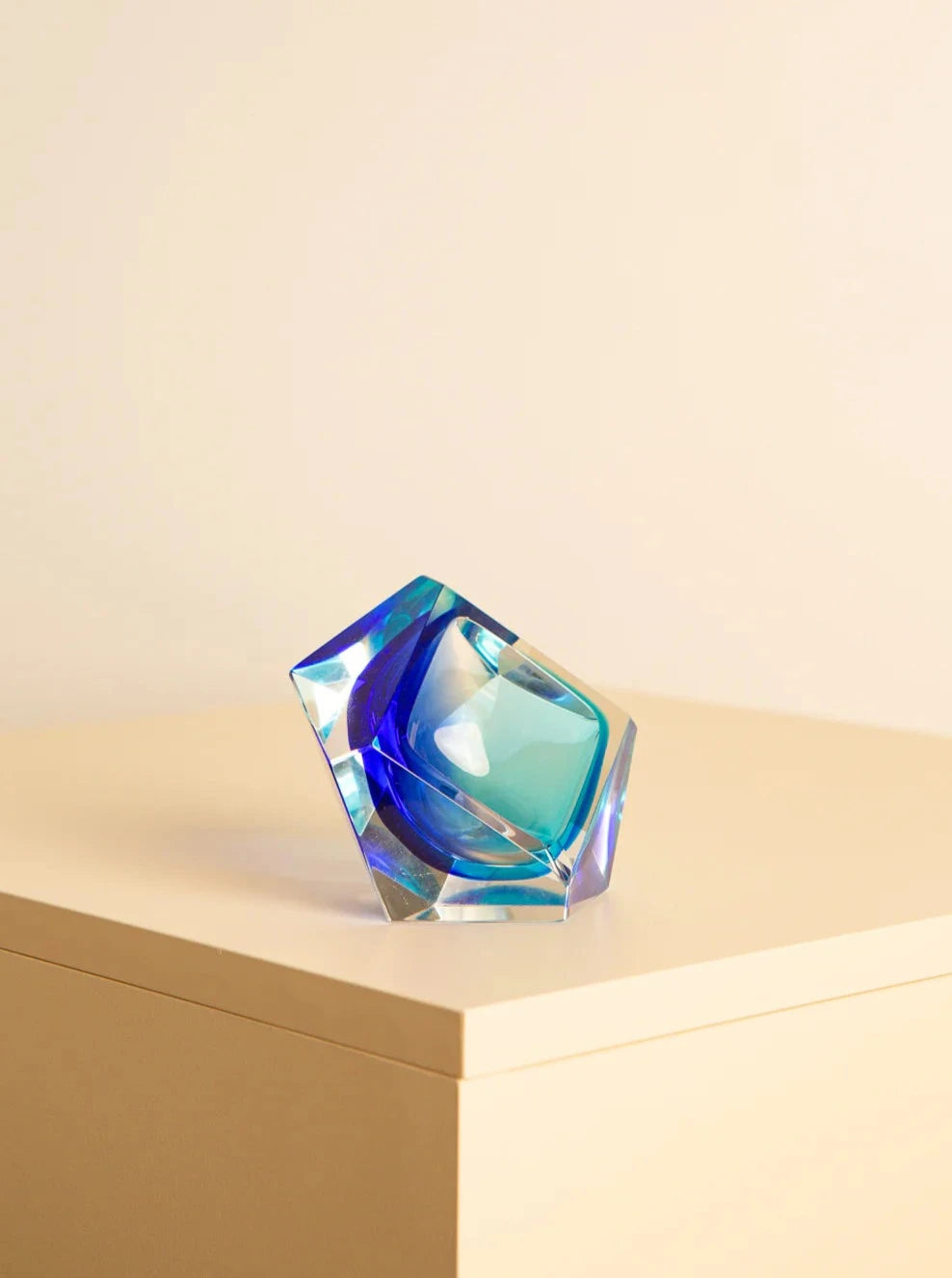 A multifaceted, iridescent Treaptyque Blue "Diamant" ashtray in murano glass by Flavio Poli for Seguso 60's with blue and green hues sits on the edge of a beige surface, angled slightly to the left. Reminiscent of Flavio Poli's Italian glass art, its colorful reflections and geometric design are emphasized by the plain, soft beige background.