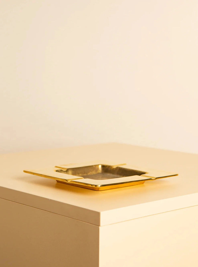 A square, gold-colored Treaptyque 60's Italian brass square ashtray with raised edges sits on a light beige surface. The piece exudes a '60s minimalist design. The background is plain and matches the color of the surface.