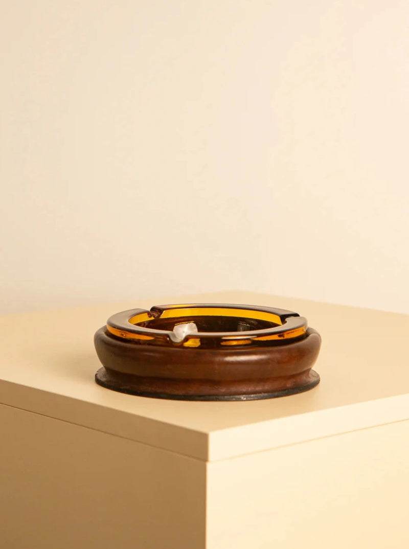 A brown glass ashtray exudes retro warmth as it sits on a light-colored, square surface against a neutral background. This circular piece with a glossy finish and a few cigarette rests along its rim could easily be mistaken for an elegant Glass and Italian leather ashtray 60's from Treaptyque.